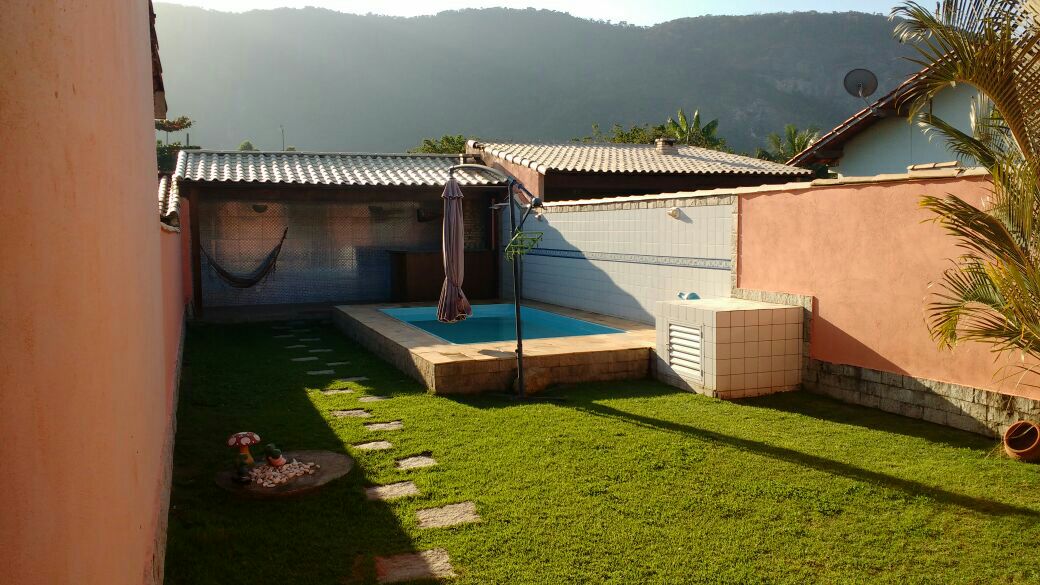A grassy outdoor area features a swimming pool surrounded by a wooden deck. A covered space is visible with a hammock, and a stone pathway leads towards a decorative fire pit. Lush green mountains can be seen in the background.