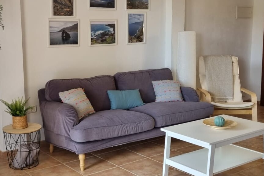 A comfortable purple sofa is complemented by decorative pillows in shades of blue and pastel colors. A small white coffee table sits in front, featuring a decorative orb. Framed landscape photos are arranged above the sofa, enhancing the living space's relaxed ambiance.
