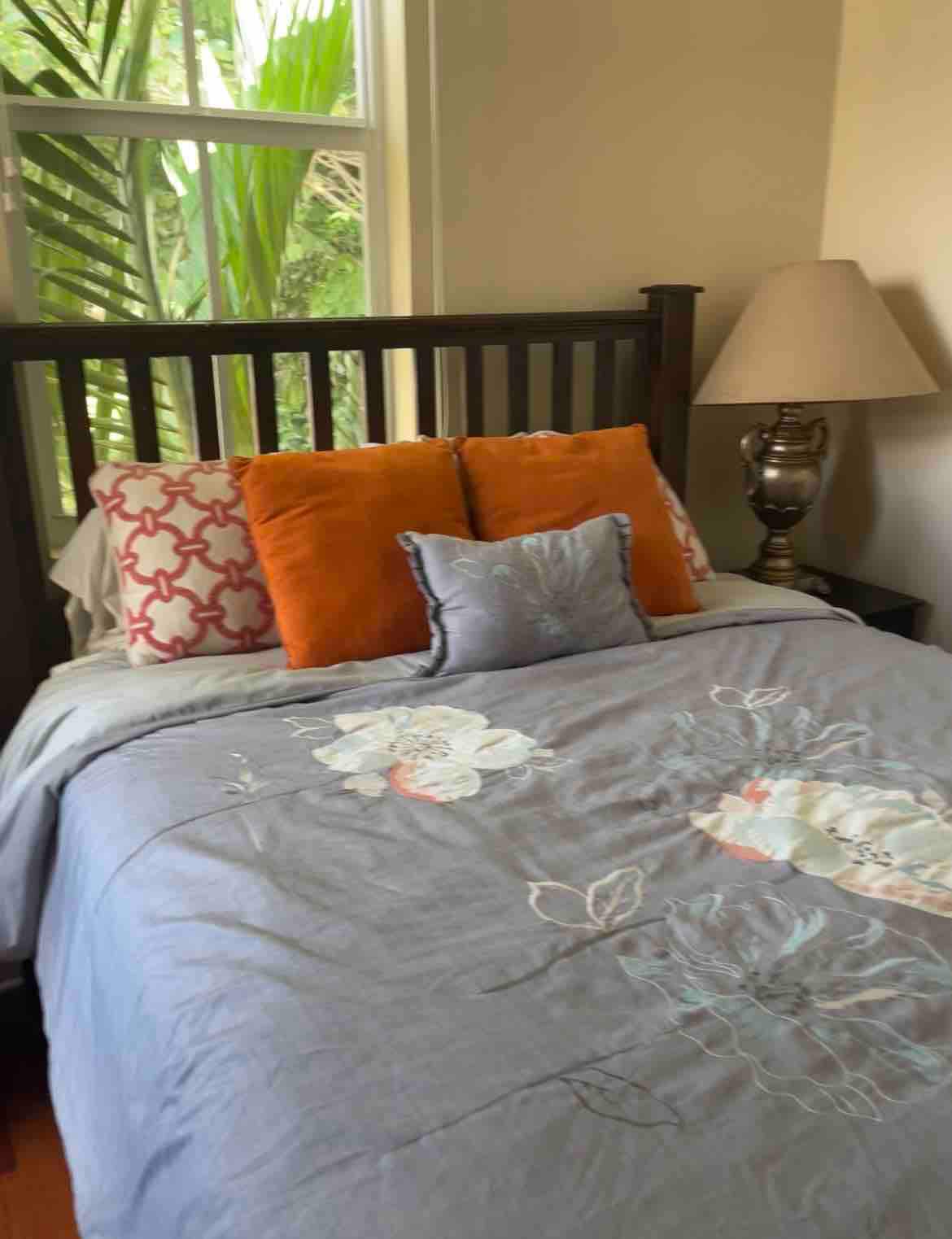 A cozy sleeping area is highlighted by a Queen-sized bed adorned with a soft, embroidered blanket featuring floral designs. Multiple decorative pillows add color and comfort. A bedside lamp provides gentle illumination, while a view of lush greenery is visible through the window.