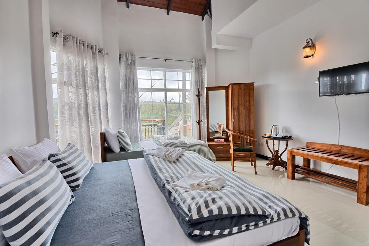 The spacious bedroom displays a large bed with striped linens, accompanied by a cozy seating area. Large windows allow natural light to fill the room, while light-colored curtains frame the view. A wooden wardrobe and a table with chairs add functionality to the elegant space.