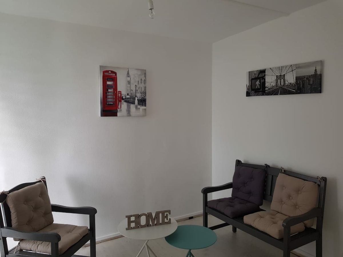 A cozy seating area is presented with two upholstered armchairs and a small circular table. Framed artwork featuring urban scenes enhances the space, while a decorative 'HOME' sign rests on the table. A light color palette creates an inviting and bright ambiance.