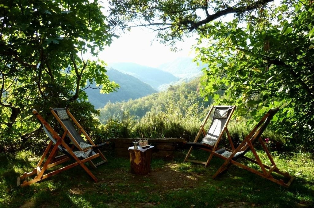 Two Adirondack chairs are positioned in a tranquil outdoor area, surrounded by greenery and trees. A small wooden table stands between the chairs, and a panoramic view of distant valleys and hills can be seen in the background, creating a peaceful atmosphere.