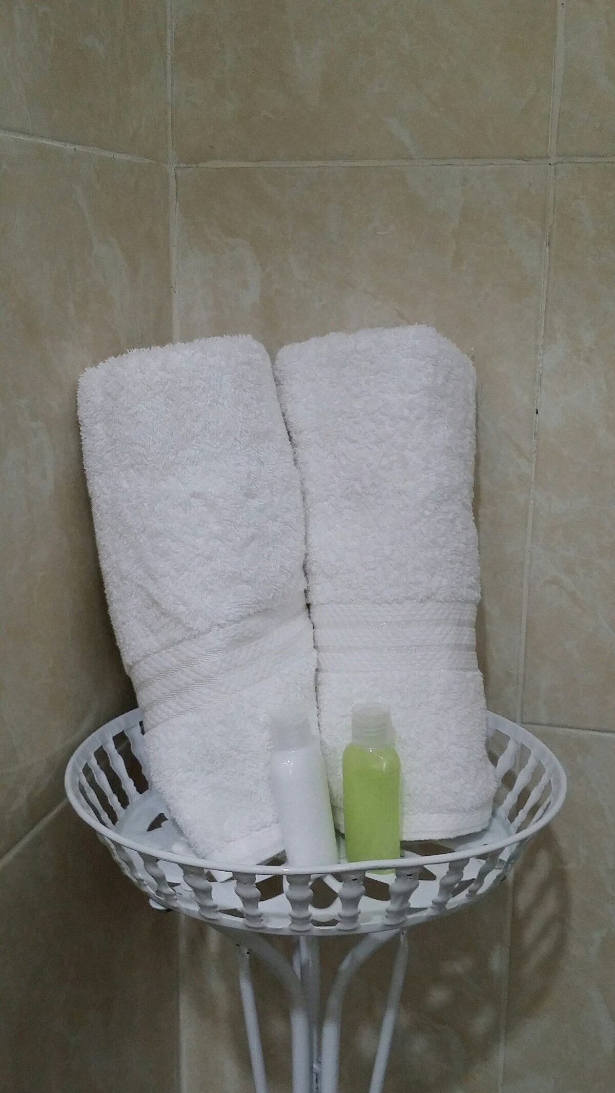 Two neatly rolled white towels are placed on a small white metal rack. Beside the towels, two bottles of toiletries are visible, one white and one green. The background features light-colored tiled walls.