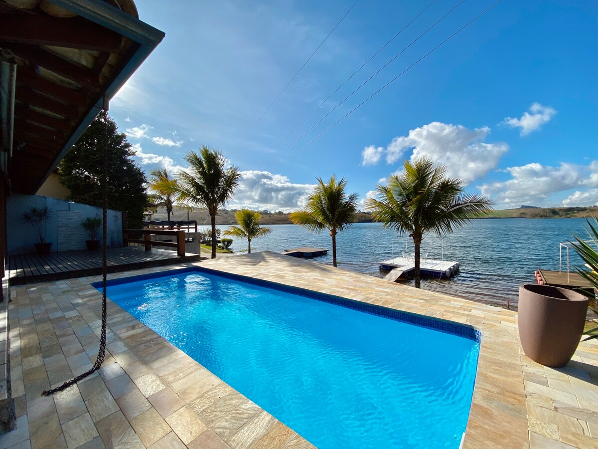 A clear blue pool rests beside a spacious patio, framed by palm trees. The water reflects the sky, which is bright with scattered clouds. A wooden deck extends toward the waterfront, offering easy access to the serene lake.