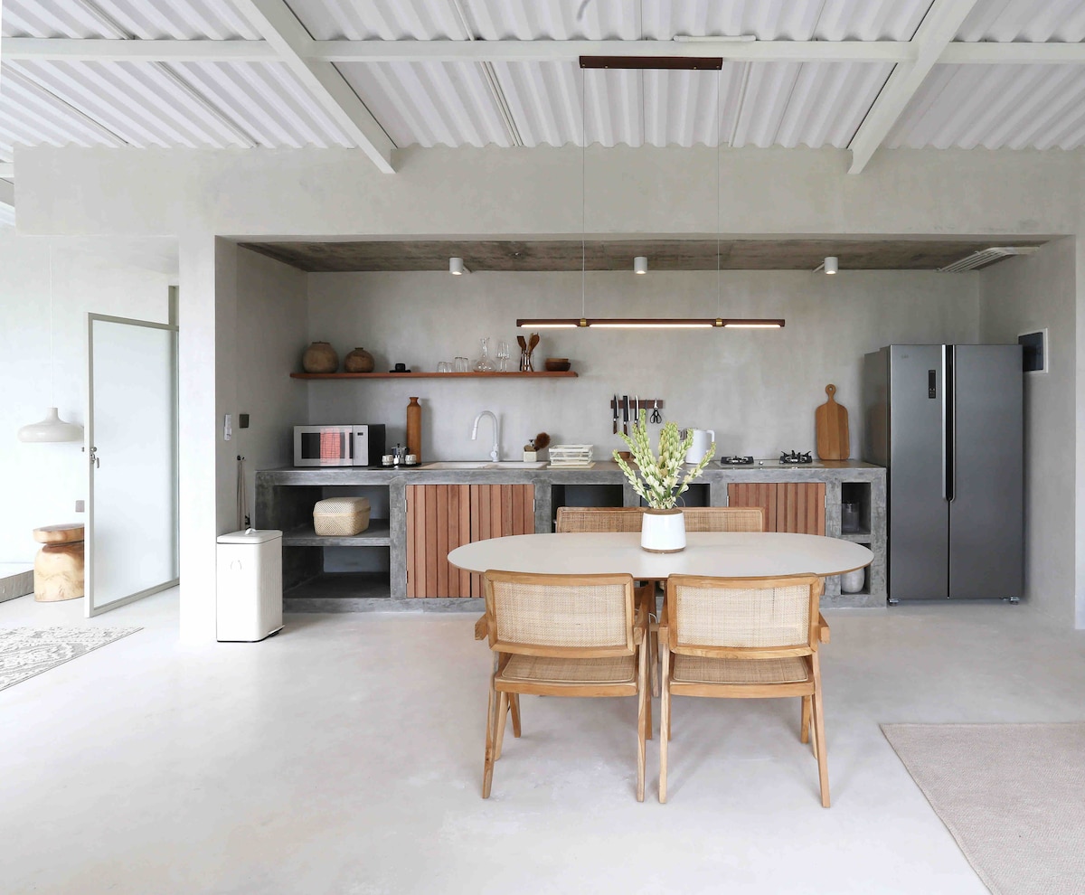 A modern kitchen is showcased with a central dining table surrounded by woven chairs. The open-concept space features concrete finishes, wooden accents, and a well-stocked kitchen counter with appliances and utensils. Natural light illuminates the area through large windows.