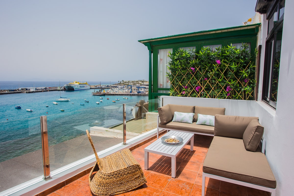 An outdoor terrace is displayed with comfortable seating arrangements, including a plush sofa and a wicker chair. The area features a glass railing, providing unobstructed views of the sea and marina. Lush green plants are visible, enhancing the tranquil setting.