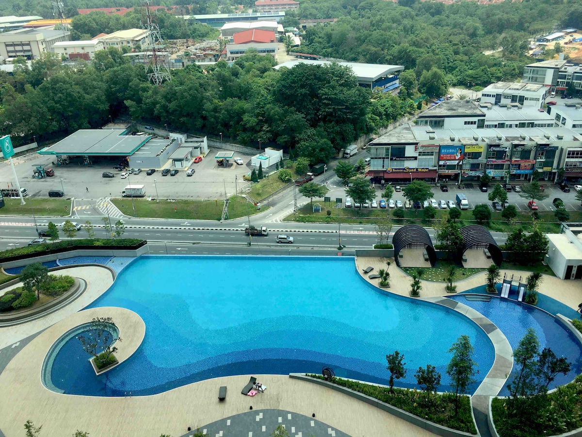 A large outdoor swimming pool is prominently displayed, featuring a curved shape and clear blue water. Surrounding greenery and well-maintained walkways complement the scene, creating a serene environment. Nearby, the road and buildings are visible in the background, offering context to the urban setting.