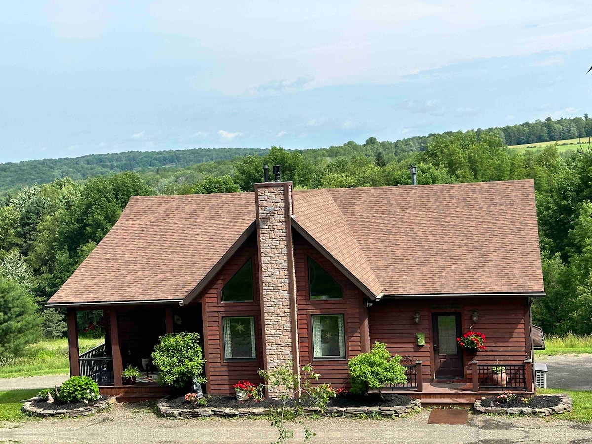 A charming country home is displayed, featuring a warm wooden exterior and a stone chimney. Bright flowers adorn the front and side, enhancing the welcoming look. Surrounding greenery and rolling hills create a picturesque backdrop, inviting guests to enjoy the peaceful outdoor environment.
