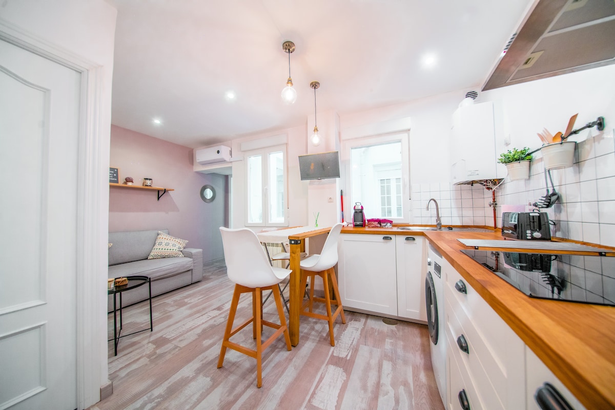 A bright and airy kitchen area features modern white cabinetry and a wooden countertop. A small dining table is surrounded by four stools. Soft light fixtures hang from the ceiling. The adjacent living area contains a light gray sofa, complemented by a wall-mounted television.