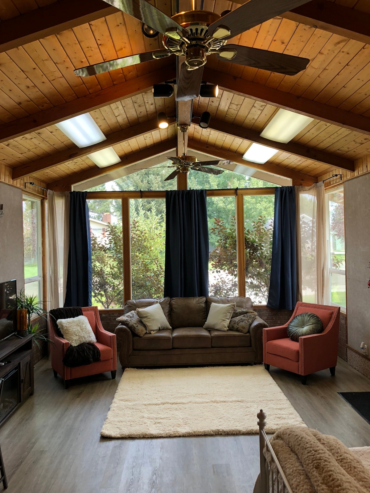 A sunlit seating area features a cozy brown sofa and two accent chairs in soft red tones. Large windows provide views of the outdoors, with sheer curtains allowing natural light to fill the space. The ceiling is adorned with exposed beams and ceiling fans.