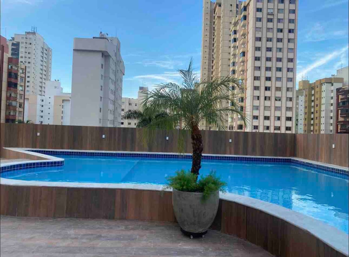 A rooftop pool area is presented, featuring clear blue water surrounded by a deck. A potted palm tree adds a touch of greenery, contrasting against the backdrop of city buildings and a clear sky.