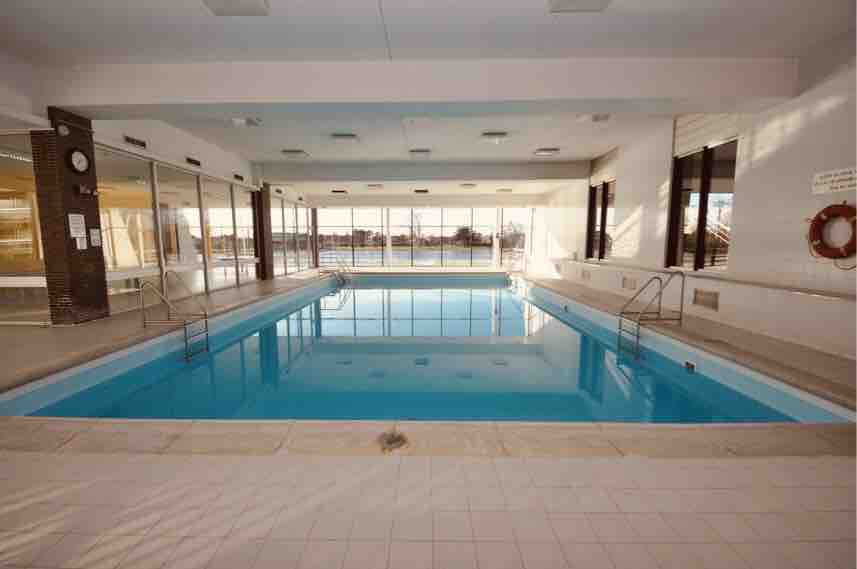 An indoor heated swimming pool is featured, surrounded by large glass windows allowing natural light to fill the space. The pool is bordered by light-colored tiles, and poolside ladder steps provide access to the water. A lifebuoy is mounted on the wall for safety.