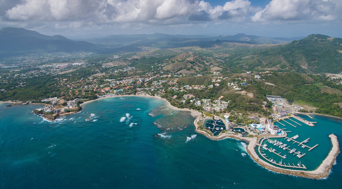 An aerial view showcases a coastal area with turquoise waters gently lapping at the shore. The landscape features a combination of lush greenery, residential areas, and a marina filled with boats. Rolling hills are visible in the background under a partly cloudy sky.