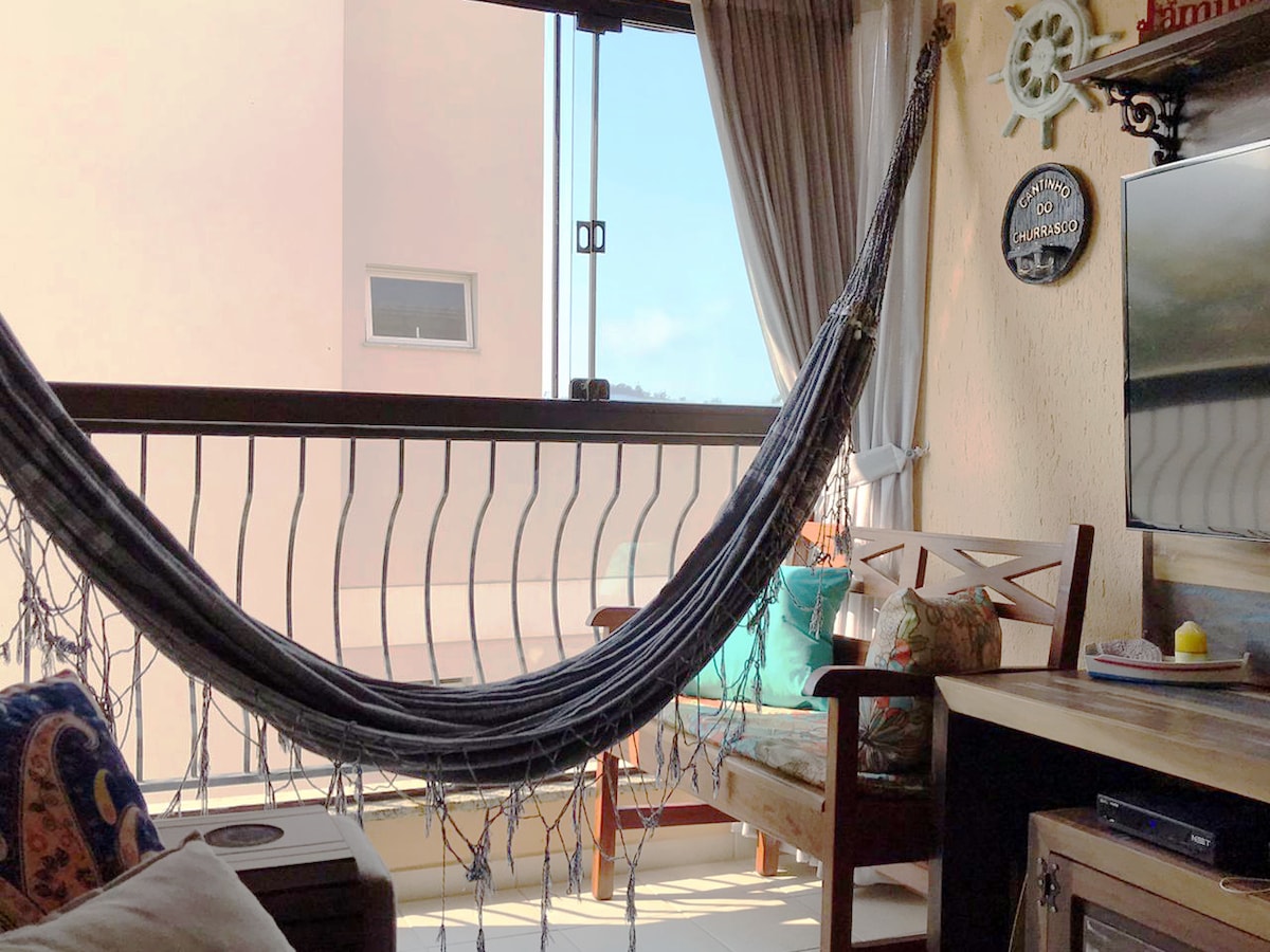 A cozy living area showcases a hammock suspended between two supports, inviting relaxation. Natural light filters through large windows, illuminating a wooden bench adorned with colorful cushions. Nearby, a television rests on a wooden cabinet, enhancing the comfort of the space.