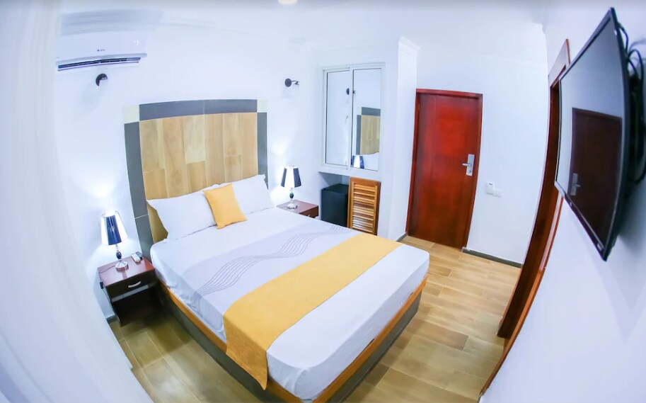 A private room features a spacious bed with a light-colored coverlet and decorative pillows. Two bedside lamps are positioned on dark wooden nightstands. A dresser and a mirror are visible, along with a wall-mounted television. Natural light enters through a nearby window, enhancing the room's brightness.