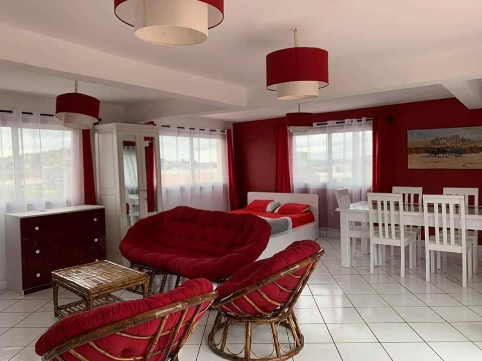 A bright living area features red accents and white furnishings. A cozy seating arrangement includes a circular red chair and wicker chairs. A dining table is set for six, complemented by large windows allowing natural light to fill the space.