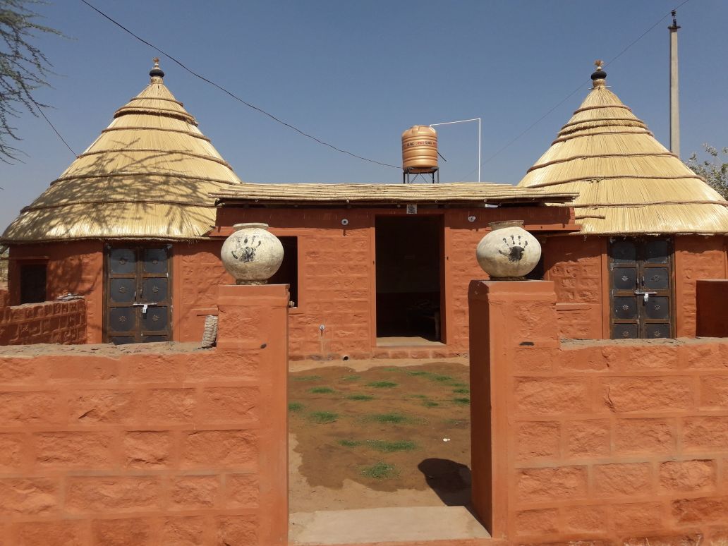 The exterior showcases a unique structure with two thatched roofs and earthen walls, framed by a low stone wall. Decorative handprints are visible on stone pots flanking the entrance, which features wooden doors and a grassy area in front.