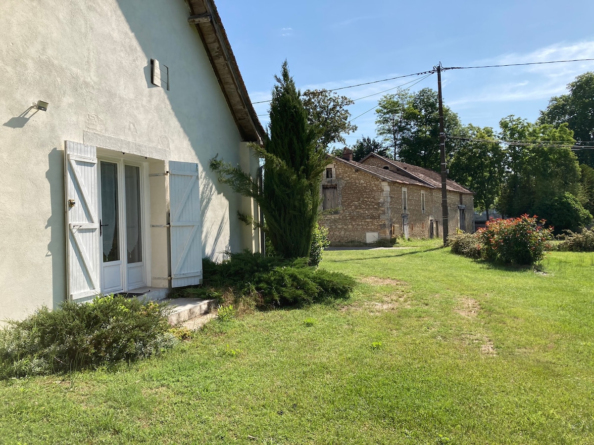 The exterior of the house features a light-colored façade with white shutters. A grassy area surrounds the building, enhanced by shrubs and a small tree. A glimpse of neighboring structures can be seen in the background, set against a clear blue sky.