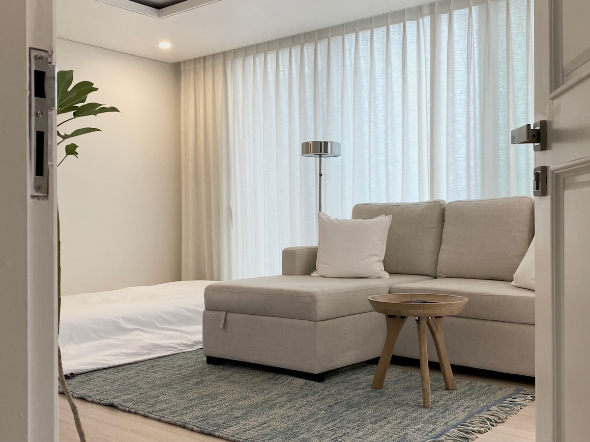 A welcoming living space is highlighted, featuring a light-colored sectional sofa with a soft cushion and a small wooden coffee table. Natural light filters through sheer curtains, and a floor lamp adds gentle illumination, creating a cozy atmosphere.