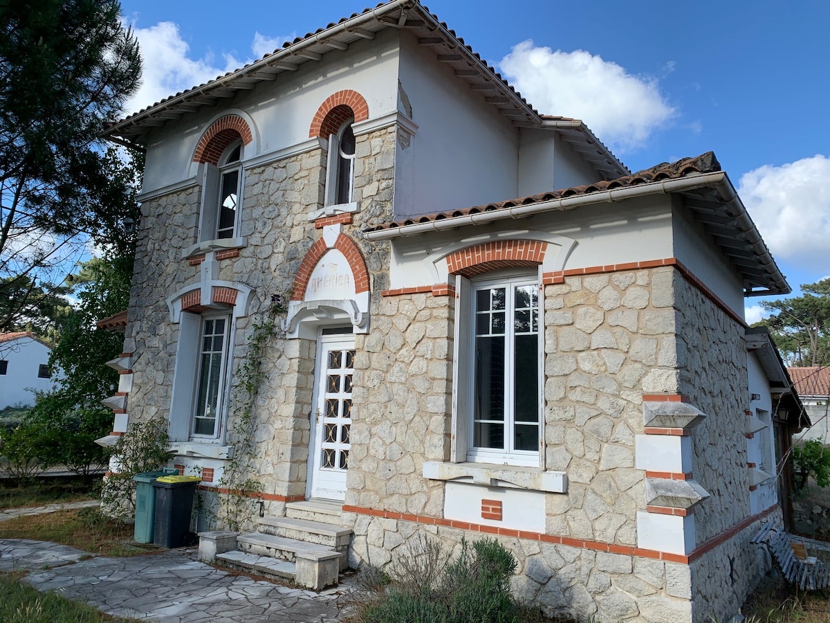 The front exterior of an elegant stone house is presented, showcasing a charming architectural style with decorative brick accents. Large windows allow natural light to fill the interior. A stone pathway leads to the entrance, bordered by greenery and a garbage bin nearby.