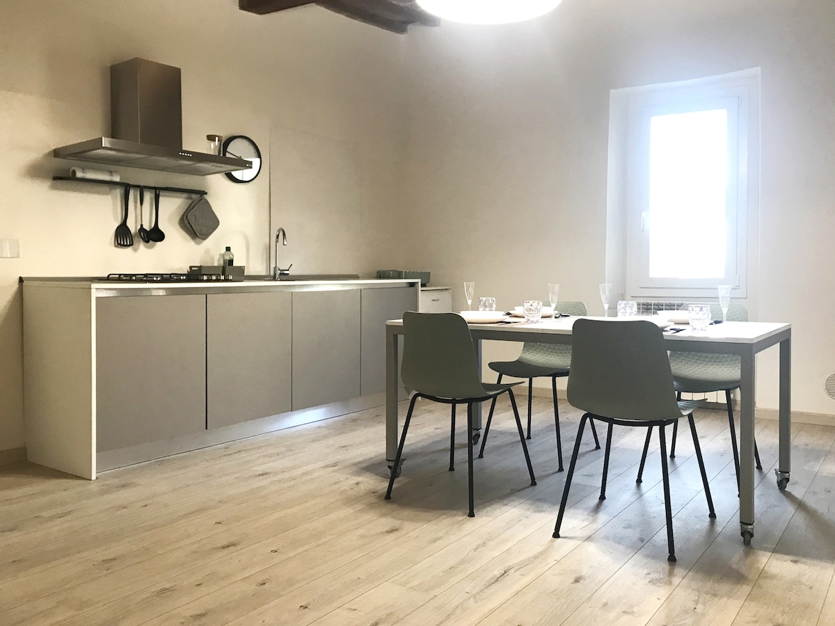 A modern kitchen area is paired with a dining space. The kitchen features sleek cabinetry and stainless steel appliances, while the dining table is surrounded by four chairs. Natural light enters through a window, highlighting the wooden flooring and neutral walls.