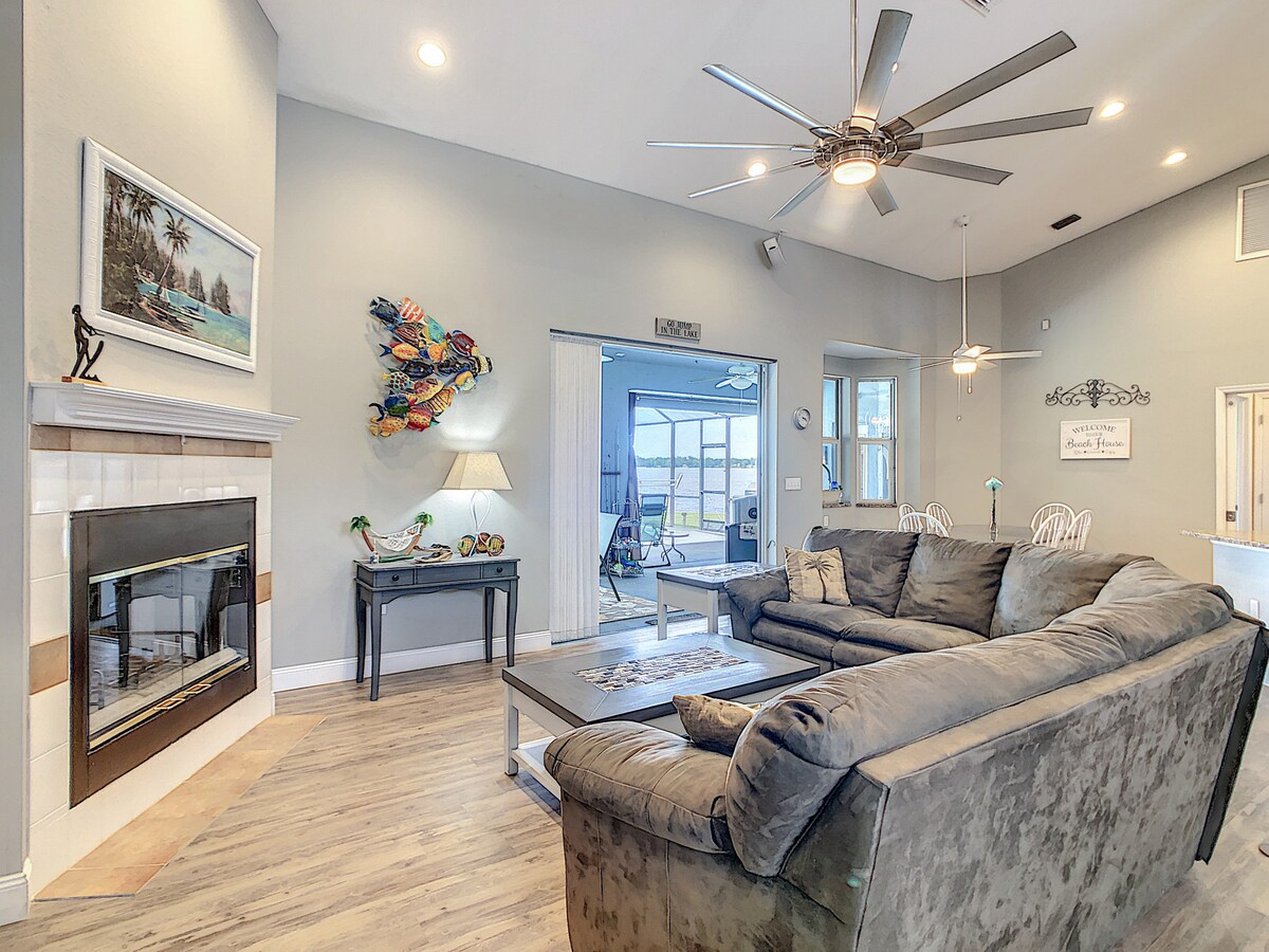 A spacious living area is centered around a plush dark sofa and a wooden coffee table. A fireplace stands against the wall, complemented by decorative artwork. Large ceiling fans circulate air above, while sliding glass doors provide access to the outdoor lanai and pool area.