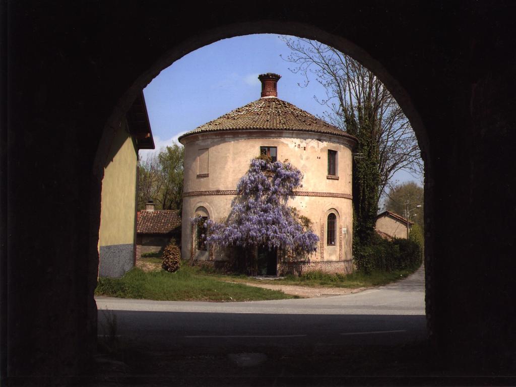 A round historical structure is framed by an archway, showcasing its unique architecture. A lush wisteria tree adds a touch of color at the front, while the surrounding greenery complements the tranquil setting. The building displays a rustic charm with a tiled roof and earthy tones.