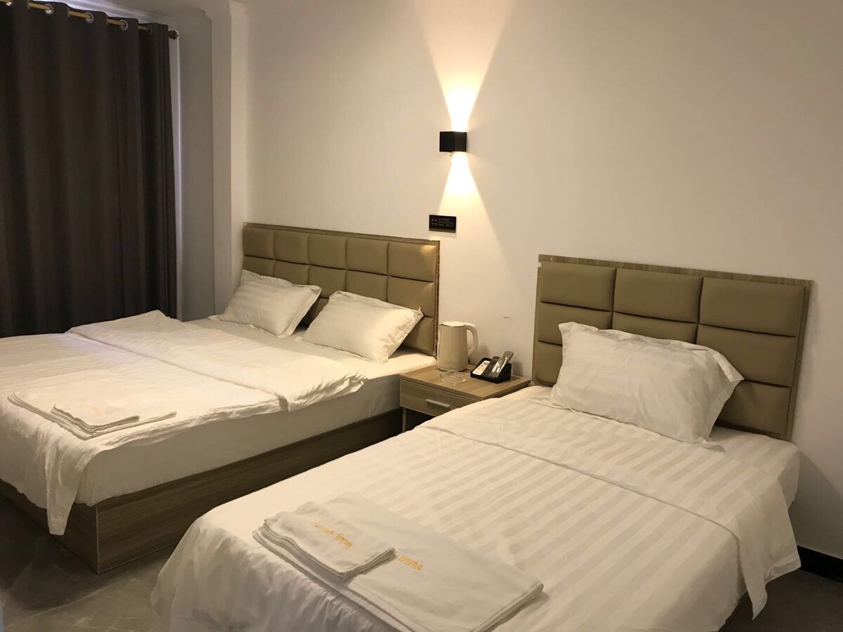 The room features two spacious beds with crisp white linens. A bedside table holds a phone and other essentials. Simple grey curtains frame the window, and soft lighting is provided by a wall-mounted fixture.