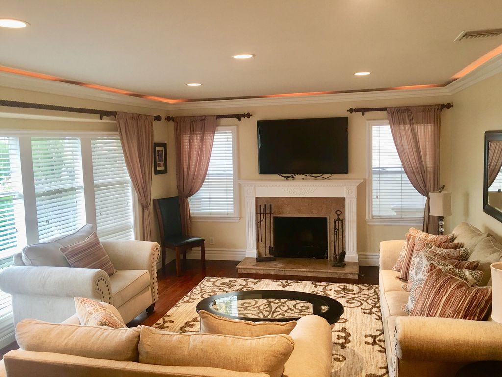 A welcoming living room features a comfortable seating arrangement with light-colored sofas and armchairs. A central coffee table is placed on a patterned area rug. Natural light filters through window treatments, highlighting the elegant fireplace and mounted television.