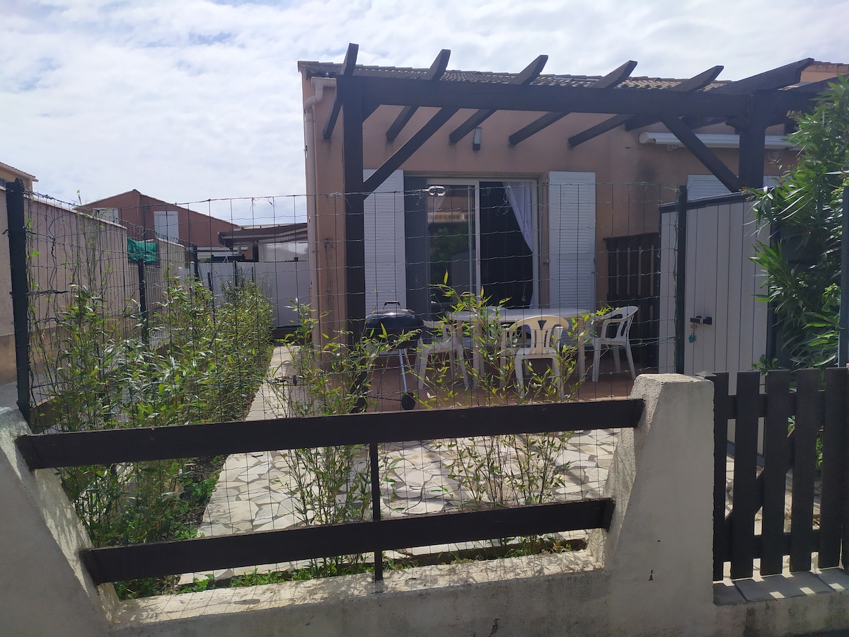 The exterior space features a fenced patio area with a stone pathway leading to a dining table and chairs under a wooden pergola. Lush greenery is present, providing a natural boundary. A small barbecue is visible, adding to the outdoor cooking possibilities.