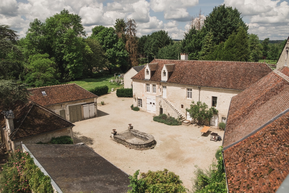 The image presents a view of the estate courtyard surrounded by lush greenery. A central stone fountain is visible, adding a touch of elegance. The rustic buildings exhibit classic architectural features, and outdoor seating areas are scattered, promoting open-air relaxation.
