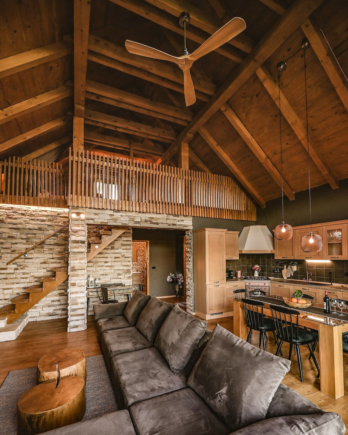A spacious living area features high wooden ceilings, with a cozy sectional sofa positioned in front of a modern kitchen. Natural materials, like exposed stone walls, accent the room. A staircase leads to an open gallery overlooking the space, enhancing the overall light and airy feel.