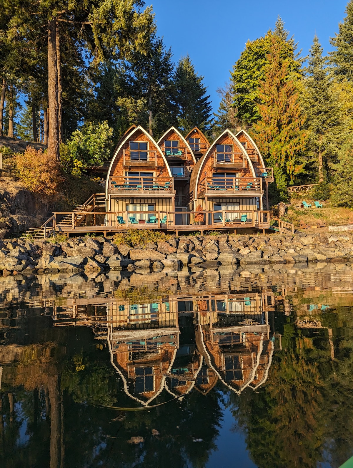 A charming waterfront building features a unique design with pointed roofs, nestled among tall trees. The structure reflects beautifully in the calm water, highlighting its rustic wooden exterior and surrounding rocky shoreline.