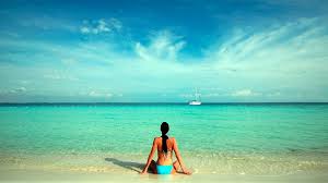 A person sits on soft sand at the water's edge, facing a tranquil sea. The water shifts from turquoise to deep blue, meeting a clear sky dotted with wispy clouds. A distant boat is visible on the horizon.