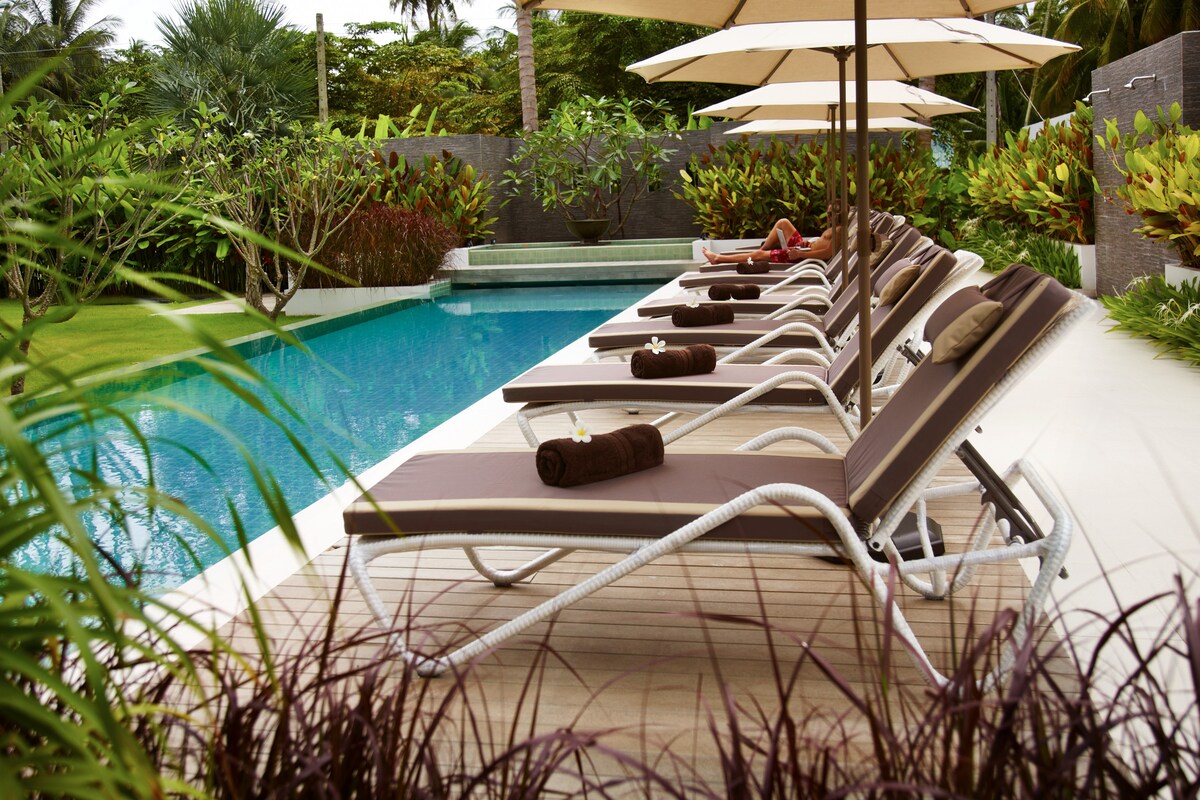 A series of sun beds are arranged neatly along a wooden deck beside a swimming pool. Each bed is equipped with a rolled towel. Lush green foliage surrounds the area, creating a soothing environment with a hint of tropical landscaping.