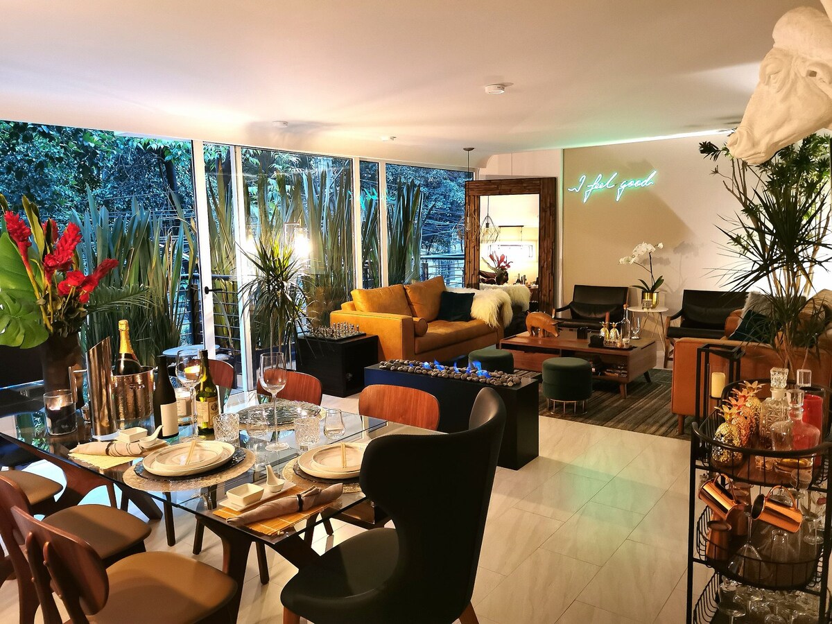 An open living and dining area showcases large windows overlooking lush greenery. A dining table is set for guests, complemented by elegant dinnerware. A cozy seating area features a mix of modern furniture, including a plush sofa and decorative plants, creating a vibrant and inviting space.