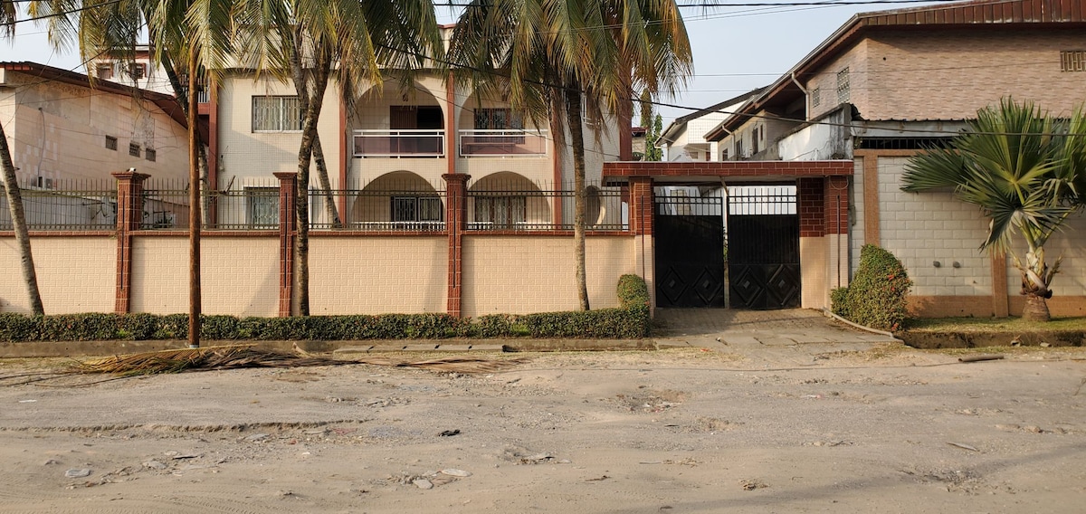A well-maintained exterior view of a two-story building featuring a gated entrance. Palm trees flank the pathway leading to the gate, and the façade includes balconies on the upper level. The surrounding garden is neatly trimmed, contributing to the overall inviting appearance.