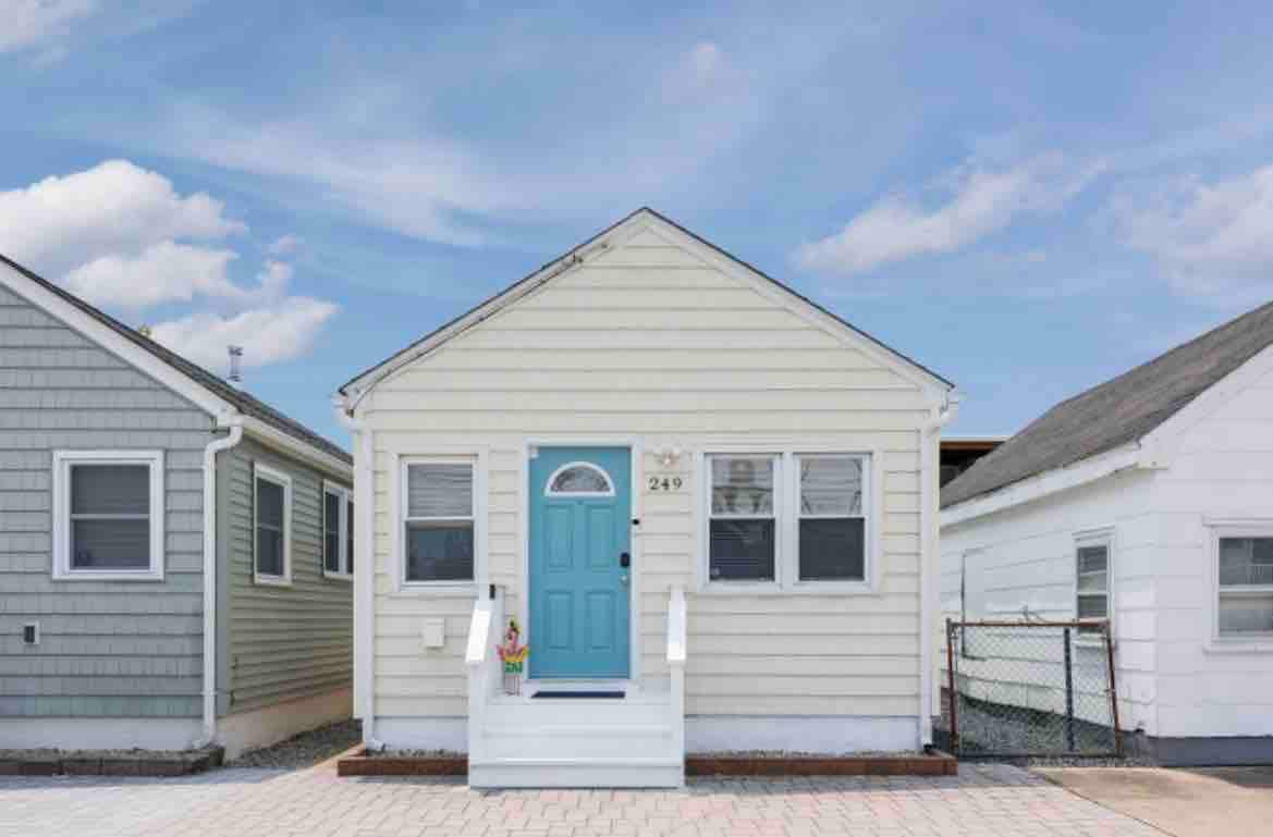 A charming beach home features a light yellow exterior with white steps leading to a blue front door. The inviting facade showcases multiple windows, providing a bright and airy appearance. Nearby structures highlight a coastal neighborhood atmosphere.