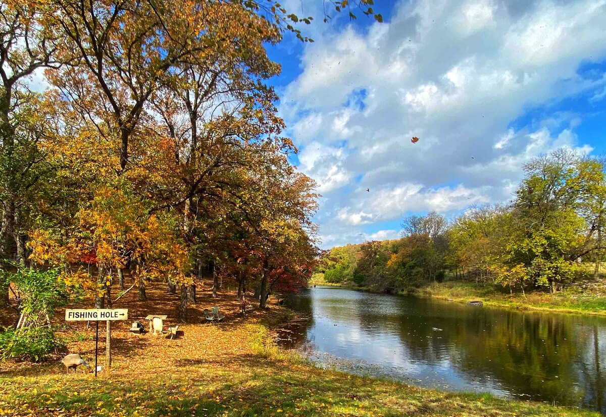 A serene fishing pond is framed by trees displaying autumn foliage. A sign reading 'Fishing Hole' is positioned along the pond's edge. Rock seating and scattered leaves create a natural, tranquil environment, reflecting a peaceful retreat in nature.