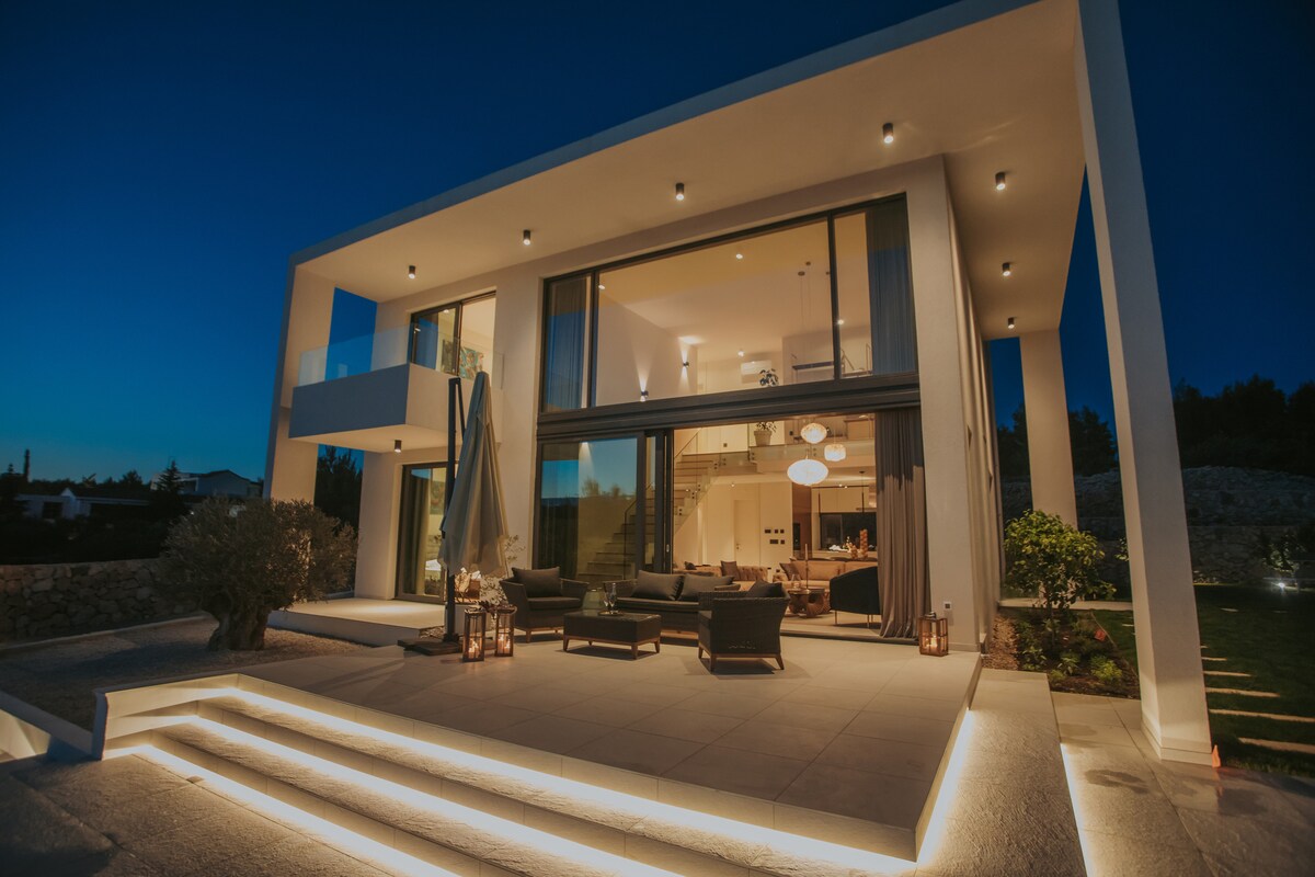 A modern villa stands illuminated at dusk, showcasing large glass doors that provide a view into the open-plan interior. Outdoor seating is arranged on the spacious terrace, with soft lighting highlighting the clean lines of the architecture and the surrounding greenery.
