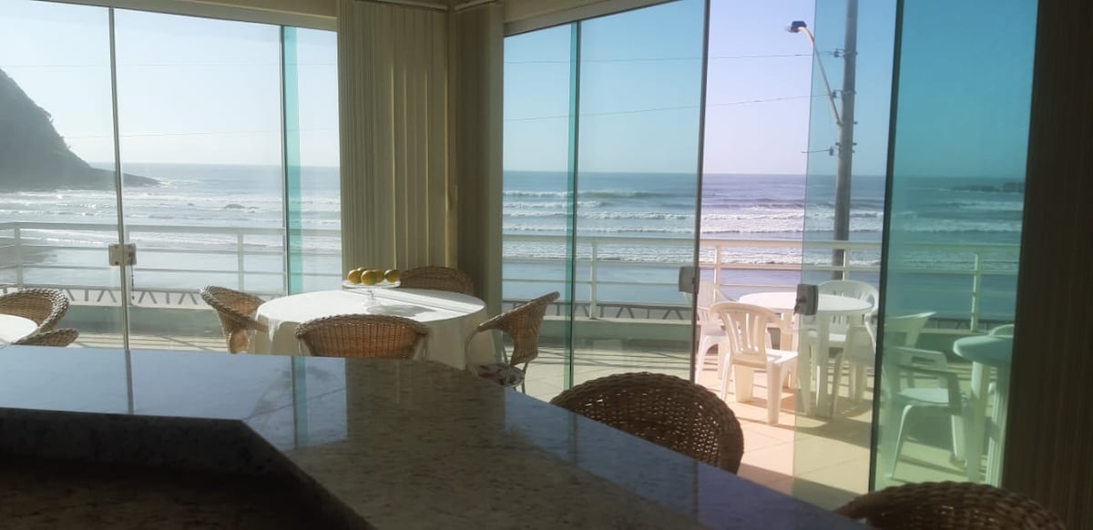 A spacious living area is framed by large glass walls, providing unobstructed views of the ocean and beach. Natural light illuminates the space, where round tables and woven chairs are arranged for dining. A countertop made of smooth stone is visible in the foreground.