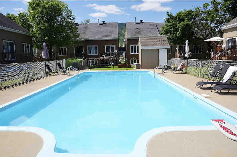 A well-maintained outdoor swimming pool is surrounded by a fenced area, featuring lounge chairs and umbrellas. The pool's clear blue water reflects the sky, while the neighboring buildings are visible in the background, providing a serene atmosphere for relaxation.