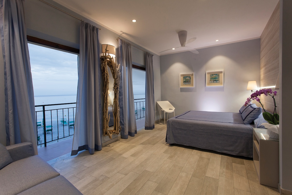 An open space suite features large windows offering sea views and inviting natural light. The room is furnished with a spacious bed, a small seating area, and soft, neutral tones throughout. Two pieces of wall art enhance the minimalist décor, complemented by light-colored curtains.