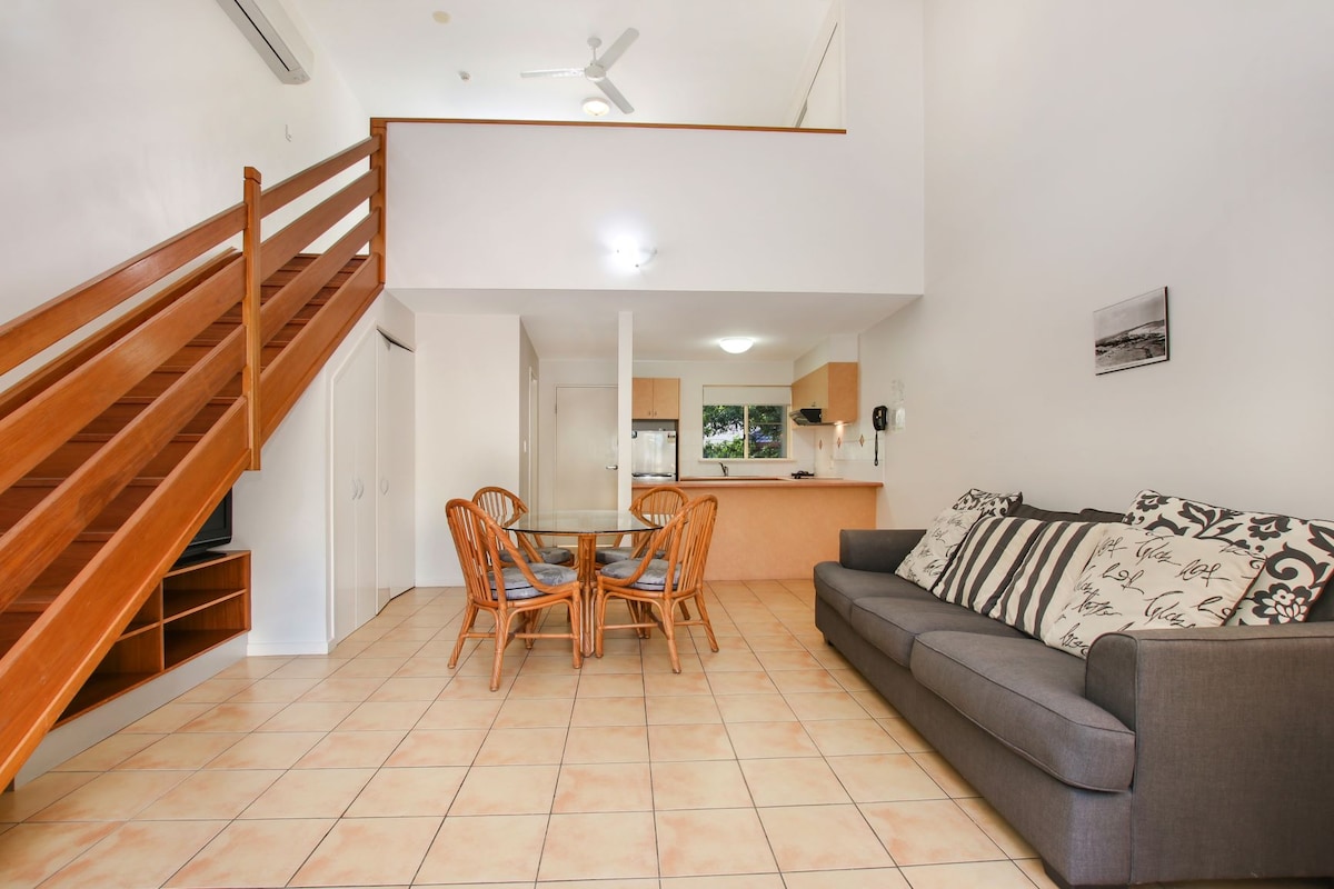 A spacious lounge dining area features a comfortable grey sofa and a round wooden dining table with four wicker chairs. The open layout reveals a staircase leading to the loft bedroom, with natural light illuminating the tiled floor.