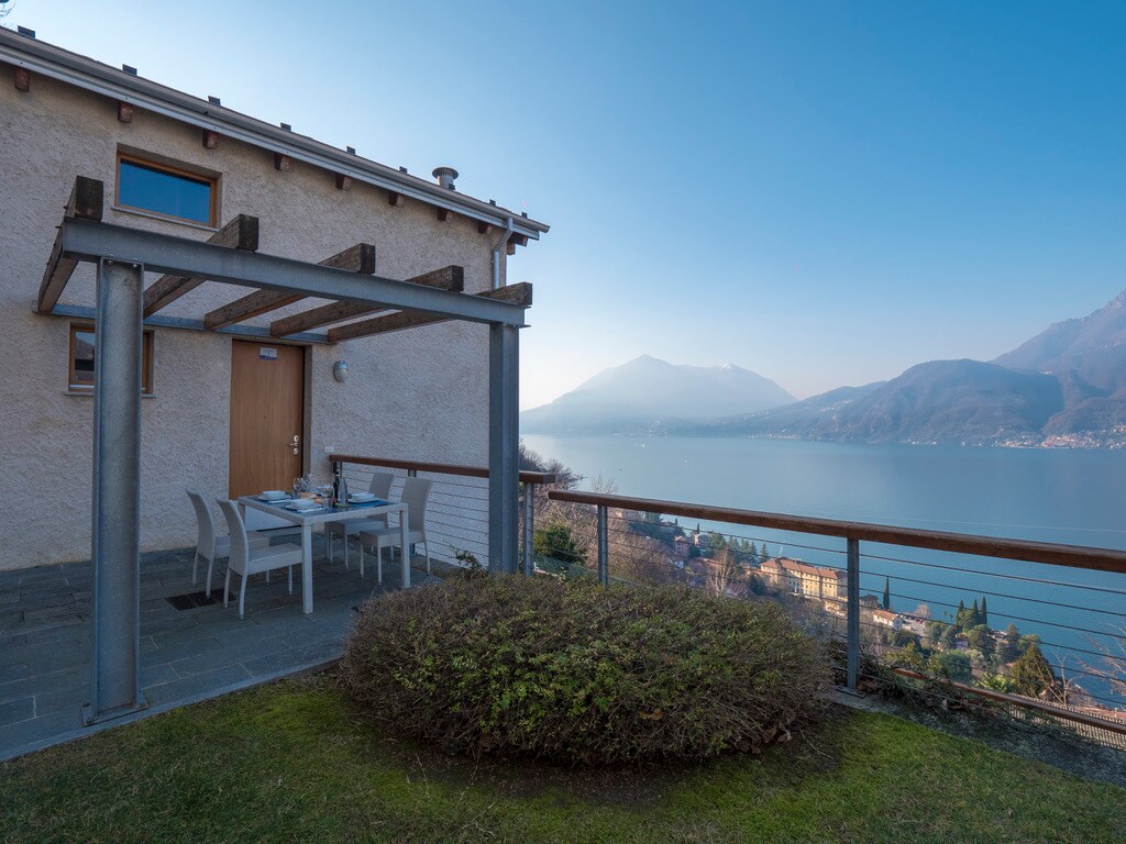 A terrace equipped with a dining table and chairs faces stunning lake views, surrounded by a well-maintained lawn. The stone pathway leads to the entrance of the duplex, framed by a wooden pergola, enhancing the outdoor experience.