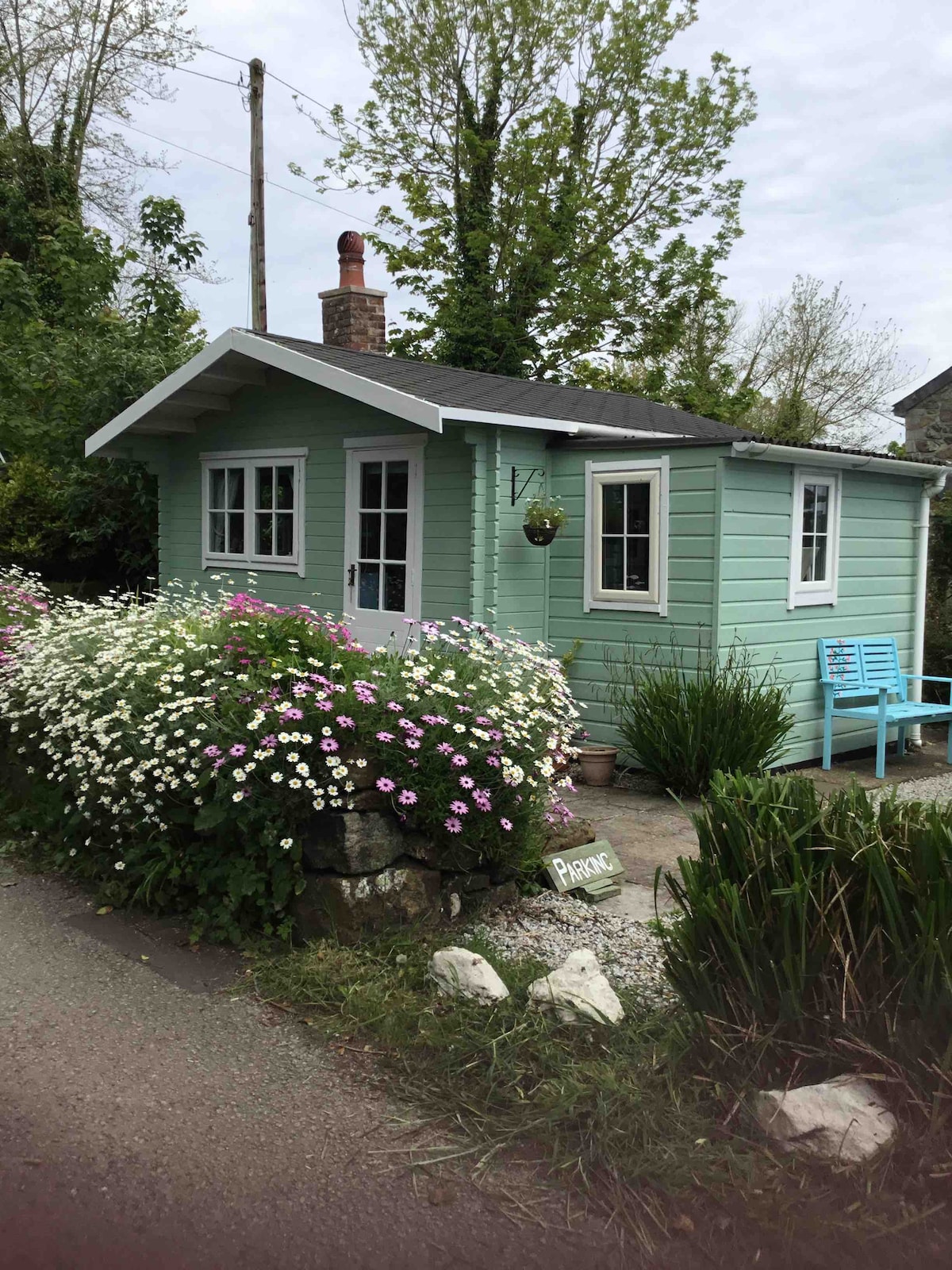 A charming cabin is situated amidst lush greenery, featuring a light green exterior and white trim. Bright flowers in a variety of colors surround the entrance, complemented by a small blue seating area. A sign is displayed nearby, indicating the name of the cabin.