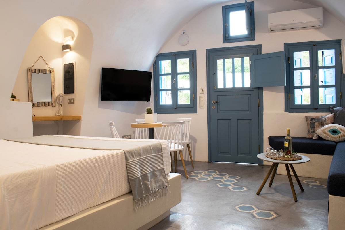 A bright and airy room features a spacious bed with neutral bedding. A seating area includes a navy sofa and a small wooden table with white chairs. Windows with blue frames allow natural light, and a mounted television adds convenience to the space.