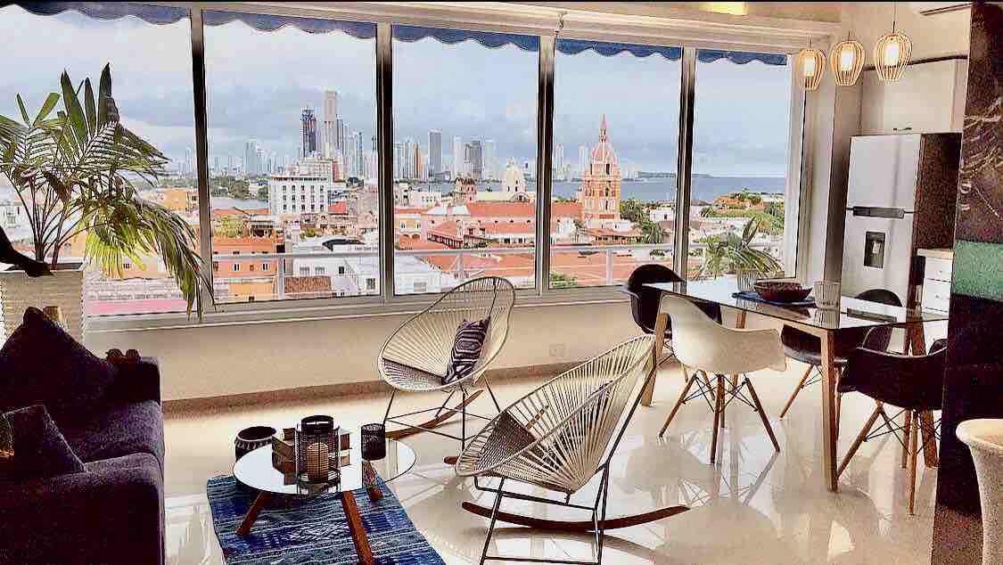 A spacious living area showcases a panoramic bay window with stunning views of the Old Town and the Caribbean Sea. Modern seating arrangements include stylish chairs around a dining table. Natural light floods the space, complemented by greenery from a nearby plant.