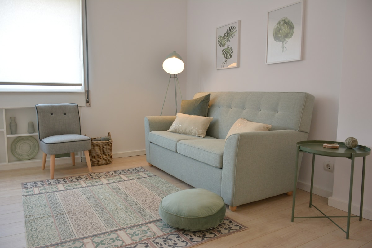 A cozy living area features a light green sofa adorned with decorative cushions, complemented by a small armchair. A round ottoman is positioned on a patterned area rug. Decorative plants and artworks are displayed on the wall and shelves, providing a serene atmosphere.
