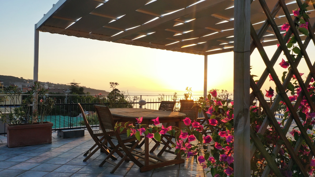 An outdoor terrace features a large wooden table surrounded by folding chairs. Pink flowers adorn the railing, complementing the scenic backdrop of a sunset over the sea. The area is illuminated by soft evening light, creating a serene atmosphere.